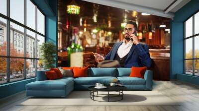 Handsome well-dressed arabian man with glass of whiskey and cigar hold mobile phone,  posed at pub. Wall mural