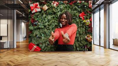 Portrait of a curly haired african woman wearing fashionable red turtleneck posing against christmas decorations, new year eve theme. Show thumbs up. Wall mural