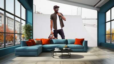 Portrait of sitting stylish african american man wear on sunglasses and cap with handbag outdoor against skate park. Street fashion black man. Wall mural