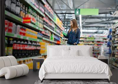 Shopping woman looking at the shelves in the supermarket.  Portrait of a young girl in a market store holding green shop basket and soft drink bottle. Wall mural