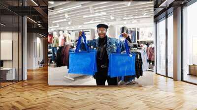 Stylish casual african american man at jeans jacket and black beret with fanny pack or waist bag holding blue shopping bags at clothes store. Wall mural