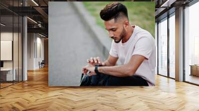 Stylish indian beard man at pink t-shirt. India model sitting at green garden of city and looking at his modern watches. Wall mural