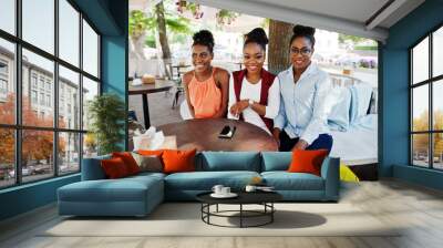 Three casual african american girls with colored shopping bags walking outdoor. Stylish black womans shopping. Wall mural