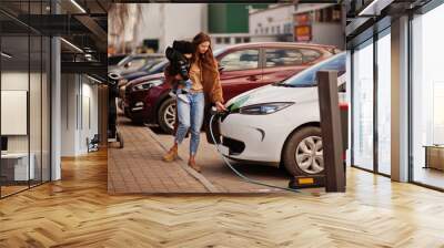 Young mother with child charging electro car at the electric gas station. Wall mural