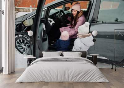 Young mother with kids charging electric car in the yard of her house at winter. Wall mural