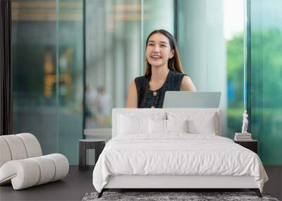 Young asian woman sitting and working on laptop indoor a cafe with big windows glasses showing greenery background Wall mural