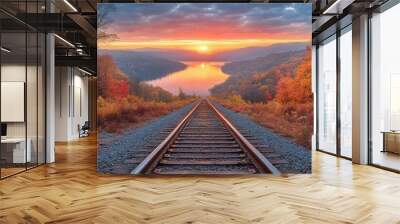 Railroad tracks lead to a distant lake surrounded by vibrant autumn foliage under a sunset sky. Wall mural