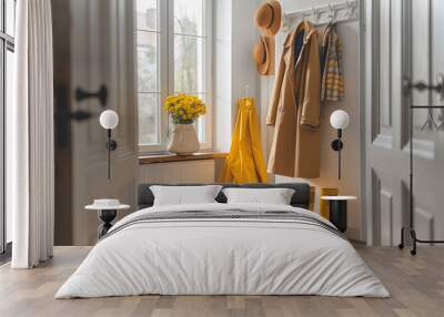 Yellow coat on a hanger and two hats in a bright hallway with rubber boots below and white vase with yellow flowers on the windowsill Wall mural