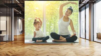 Asian mom practice yoga at home with a adorable daughter sitting next to her, trying to imitate the mother's posture with a smiling face while exercise at home. Wall mural