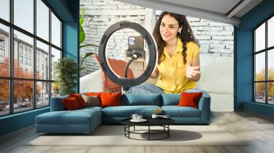 A young Caucasian influencer, creatively recording a vlog on her sofa with a ring light and mobile, sharing inspiration and connecting online. Wall mural