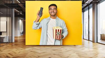 Asian man with popcorn and TV remote, embodying a cinema concept on a yellow background. Wall mural