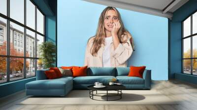 Blonde young caucasian woman in blue studio biting fingernails, nervous and very anxious. Wall mural