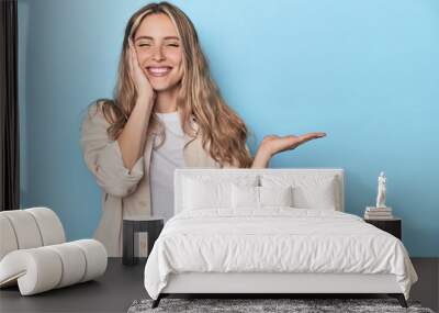 Blonde young caucasian woman in blue studio holds copy space on a palm, keep hand over cheek. Amazed and delighted. Wall mural