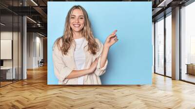 Blonde young caucasian woman in blue studio smiling cheerfully pointing with forefinger away. Wall mural