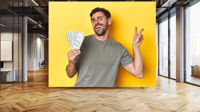 Caucasian man holding dollars, yellow studio shot joyful and carefree showing a peace symbol with fingers. Wall mural