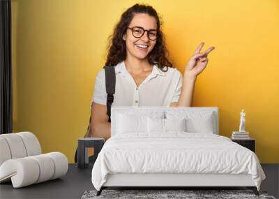 Caucasian university student with glasses, backpack, joyful and carefree showing a peace symbol with fingers. Wall mural