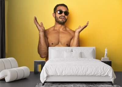 Fit young Latino man in swimwear and sunglasses, yellow studio background, surprised and shocked. Wall mural