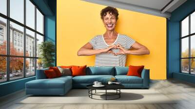 Mid-aged caucasian woman on vibrant yellow smiling and showing a heart shape with hands. Wall mural