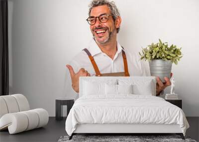 Middle age gardener caucasian man holding a plant isolated on white background  points with thumb finger away, laughing and carefree. Wall mural