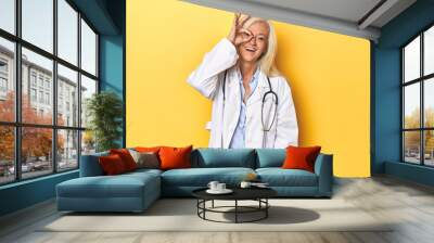 Middle-aged Caucasian female doctor on yellow studio backdrop Middle-aged Caucasian female doctor on yellow studio backdropexcited keeping ok gesture on eye. Wall mural