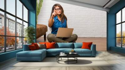 Portrait of young latin woman sitting on the floor showing number one, symbol of counting, concept of mathematics, confident and cheerful. Holding a laptop. Wall mural