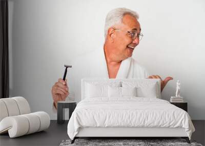 Senior american man wearing bathrobe holding razor blade isolated on white background points with thumb finger away, laughing and carefree. Wall mural