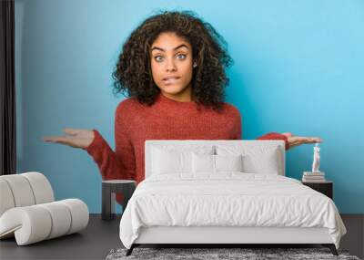 Young african american curly hair woman confused and doubtful shrugging shoulders to hold a copy space. Wall mural