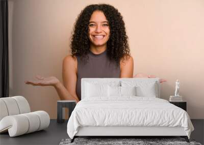 Young african american curly woman isolated makes scale with arms, feels happy and confident. Wall mural