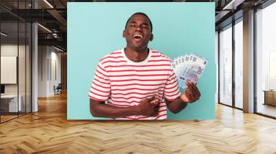 Young African American man holding a bank notes isolated on blue background laughing and having fun. Wall mural