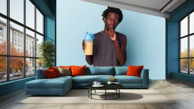 Young african american man holding protein milkshake isolated on blue background looking sideways with doubtful and skeptical expression. Wall mural