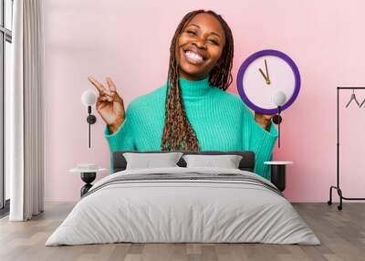 Young African American woman holding a clock isolated on pink background joyful and carefree showing a peace symbol with fingers. Wall mural