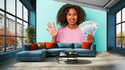 Young african american woman holding euro banknotes isolated on blue background smiling cheerful showing number five with fingers. Wall mural