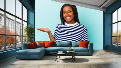 Young african american woman isolated on blue background  showing a copy space on a palm and holding another hand on waist. Wall mural