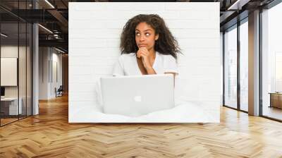 Young african american woman sitting on the bed with laptop looking sideways with doubtful and skeptical expression. Wall mural