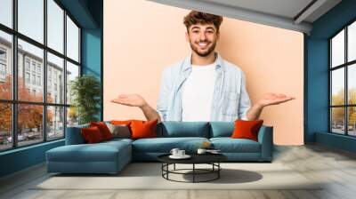 Young arab man isolated on beige background makes scale with arms, feels happy and confident. Wall mural