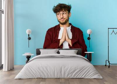 Young arab man on blue background praying, showing devotion, religious person looking for divine inspiration. Wall mural