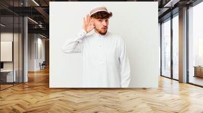 Young Arab man wearing the typical arabic costume isolated on white background trying to listening a gossip. Wall mural