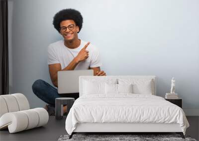 Young black man sitting on the floor with a laptop smiling and pointing to the side Wall mural