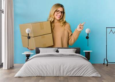 Young blonde caucasian woman holding boxes to move smiling and pointing aside, showing something at blank space. Wall mural