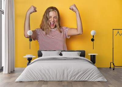 Young blonde Caucasian woman in a red striped t-shirt on a yellow background, raising fist after a victory, winner concept. Wall mural