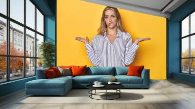 Young blonde Caucasian woman in a striped business shirt on a yellow background, confused and doubtful shrugging shoulders to hold a copy space. Wall mural