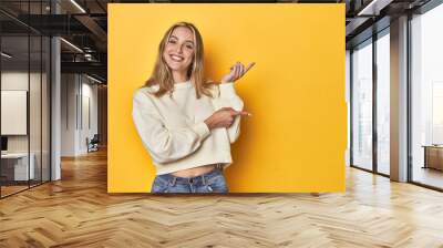 Young blonde Caucasian woman in a white sweatshirt on a yellow studio background, excited pointing with forefingers away. Wall mural