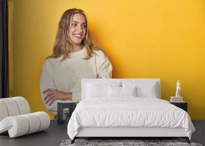 Young blonde Caucasian woman in a white sweatshirt on a yellow studio background, smiling confident with crossed arms. Wall mural