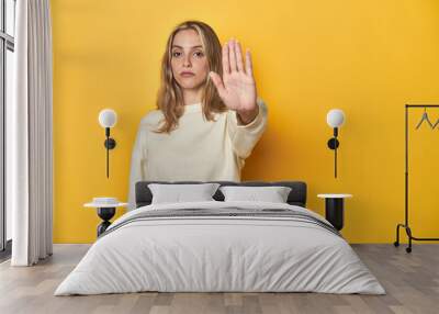 Young blonde Caucasian woman in a white sweatshirt on a yellow studio background, standing with outstretched hand showing stop sign, preventing you. Wall mural