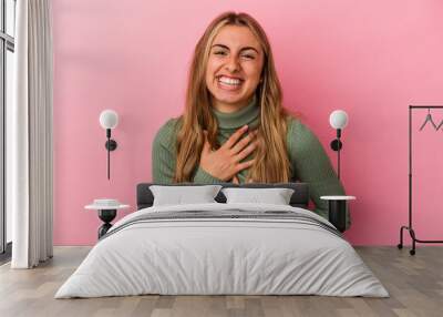 Young blonde caucasian woman isolated on pink background laughs happily and has fun keeping hands on stomach. Wall mural