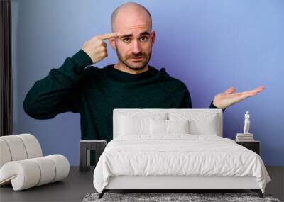 Young caucasian bald man isolated on purple background holding and showing a product on hand. Wall mural