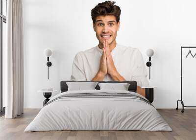 Young caucasian handsome man isolated holding hands in pray near mouth, feels confident. Wall mural