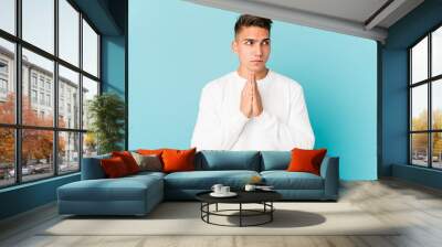 Young caucasian handsome man isolated praying, showing devotion, religious person looking for divine inspiration. Wall mural