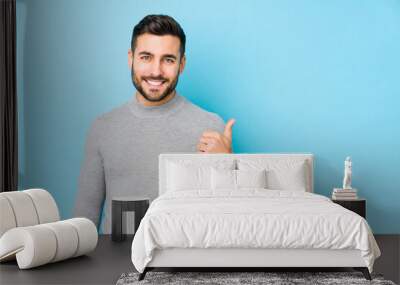 Young caucasian man against a blue background isolated smiling and raising thumb up Wall mural