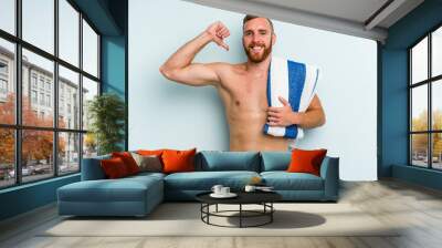 Young caucasian man going to the beach holding a towel isolated on blue background feels proud and self confident, example to follow. Wall mural